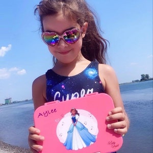 May include: A pink lunchbox with a princess illustration and the name "Aylee" written on it. The lunchbox has a white background with musical notes and a heart.