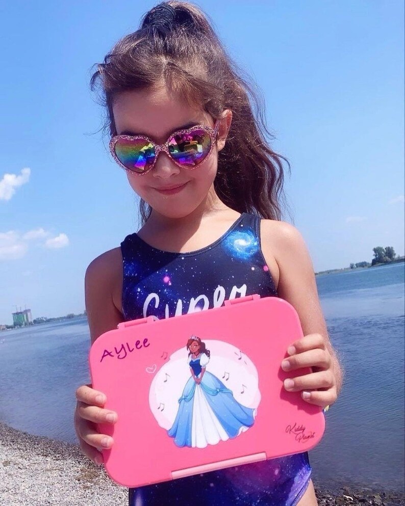 May include: A pink lunchbox with a princess illustration and the name "Aylee" written on it. The lunchbox has a white background with musical notes and a heart.