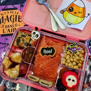 May include: A pink lunchbox with a variety of Halloween-themed snacks, including chips, fruit, and candy. The lunchbox has a cute cartoon candy corn character on the lid. The text "Boo!" is on a black label.