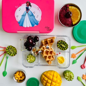 May include: A pink lunchbox with a cartoon princess on the lid, a clear lunch container with compartments filled with food, including a waffle, fruit, and a hard-boiled egg, and a green lidded container with goldfish crackers.