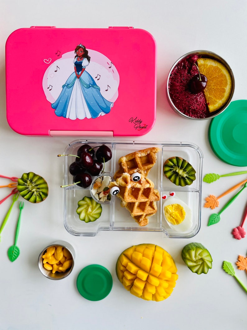 May include: A pink lunchbox with a cartoon princess on the lid, a clear lunch container with compartments filled with food, including a waffle, fruit, and a hard-boiled egg, and a green lidded container with goldfish crackers.