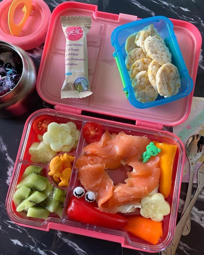 May include: Pink lunchbox with a variety of foods, including salmon, kiwi, cucumber, bell peppers, and rice cakes. The lunchbox also includes a small package of chocolate chip granola bars.