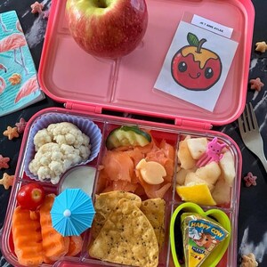 May include: Pink lunchbox with compartments filled with a variety of foods, including a red apple, cauliflower, salmon, chips, fruit, and cheese. The lunchbox has a sticker that says "Je t'aime Ayles."