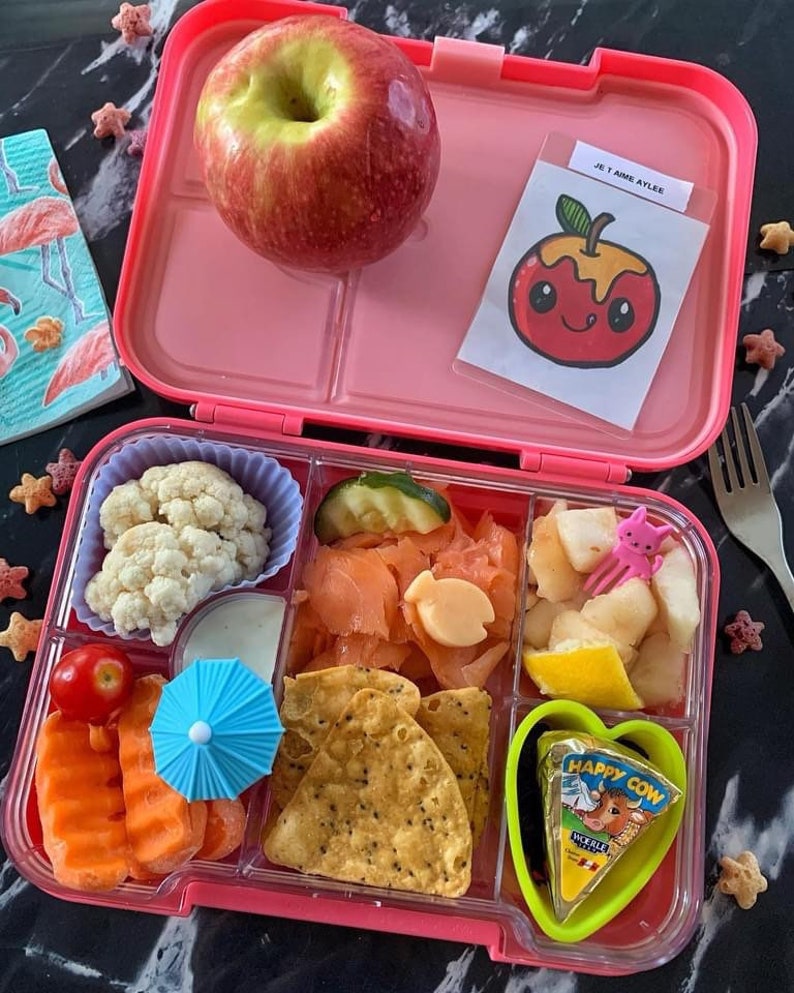 May include: Pink lunchbox with compartments filled with a variety of foods, including a red apple, cauliflower, salmon, chips, fruit, and cheese. The lunchbox has a sticker that says "Je t'aime Ayles."