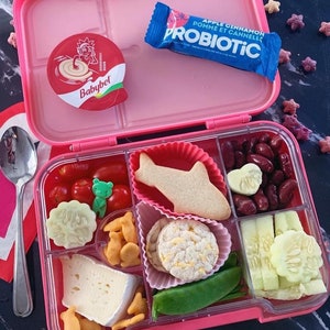 May include: A pink lunchbox with a variety of snacks inside. The lunchbox has a clear lid and is divided into multiple compartments. The snacks include a fish-shaped cookie, a small container of red beans, a small container of cherry tomatoes, a small container of cucumber slices, a small container of green peas, a small container of cheese, a small container of crackers, and a small container of rice cakes. The lunchbox also includes a small container of apple cinnamon probiotic.