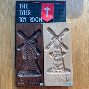 May include: Two wooden cookie cutters shaped like windmills. The cutter on the left is dark brown and says "Wipmolen 1". The cutter on the right is light brown and says "Steltingmaker 2".