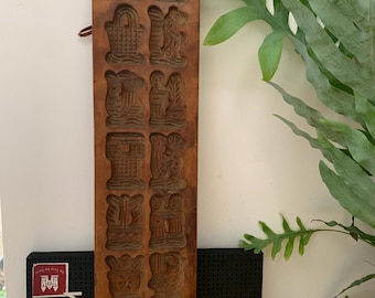Op de afbeelding: Een vintage, rechthoekige houten mal met gebeeldhouwde reliëfontwerpen. De mal bevat verschillende gedetailleerde afbeeldingen, waaronder dieren en figuren, gerangschikt in rijen. Het hout heeft een warme, bruine tint en een kleine metalen lus is aan de zijkant bevestigd. De mal wordt waarschijnlijk gebruikt voor het maken van decoratieve items.