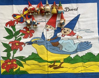 May include: A white and blue decorative cloth featuring a whimsical illustration of a gnome riding a bird, with a butterfly and flowers. Several gnome figurines are arranged above the illustration. The name "David" is written in cursive.