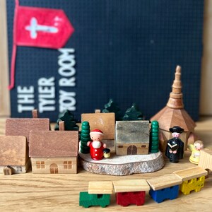 May include: Wooden toy village scene featuring miniature houses, trees, a church, and figures. The scene includes a Santa Claus figure and a small train. A banner in the background reads "THE TYLER TOY ROOM".