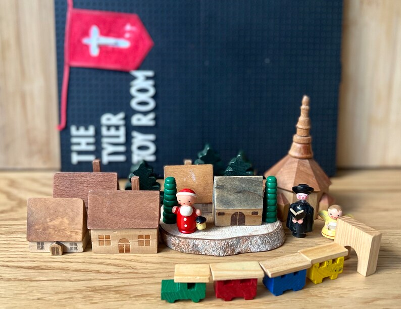 May include: Wooden toy village scene featuring miniature houses, trees, a church, and figures. The scene includes a Santa Claus figure and a small train. A banner in the background reads "THE TYLER TOY ROOM".
