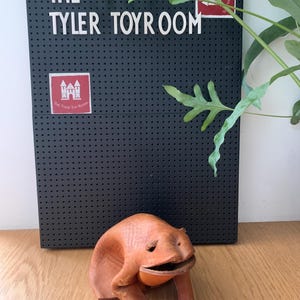 May include: A handmade, orange-brown leather frog ornament with an open mouth and outstretched legs. The frog is in front of a black pegboard sign that reads "THE TYLER TOYROOM". Green leaves are in the background.