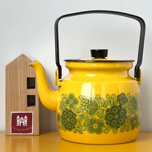 Puede incluir: Una tetera de esmalte amarillo con asa y tapa negras. La tetera tiene un patrón floral verde alrededor de la parte inferior.