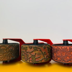 May include: Three vintage red and black rubber stamp rollers with intricate designs. The rollers are mounted on a red plastic base with a handle.