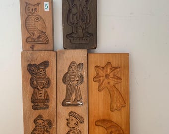 May include: A collection of vintage wooden cookie molds. The molds feature carved designs of an owl, a man, a star with a comet tail, a crescent moon, and other figures. The molds are numbered and made of dark wood.
