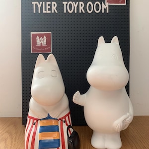 May include: Two white ceramic Moomin figures. One figure is wearing a red and white striped apron with blue and orange accents and a black bag. The other figure is holding a small object. The background features a sign that reads "THE TYLER TOYROOM".