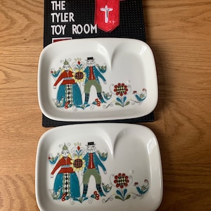 May include: Two white ceramic plates with a blue and green floral design featuring a couple in traditional folk costumes. The plates have a heart-shaped indentation in the center.