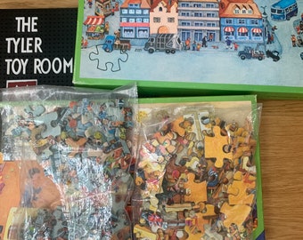 Può includere: Un puzzle colorato che rappresenta una piazza cittadina con edifici, persone e auto. Il puzzle è in una scatola con un bordo verde e le parole "The Tyler Toy Room" sul lato. I pezzi del puzzle sono in sacchetti di plastica.