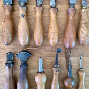 Op de afbeelding: Een verzameling van twaalf vintage lederbewerkingsgereedschappen met gladde, gedraaide houten handvatten en diverse metalen koppen, waaronder ritsers, kantenversnijders, stikwieltjes en priemen, gerangschikt in twee rijen op een vlak.