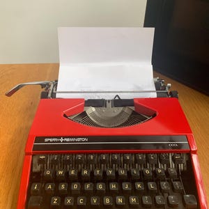 May include: A white ceramic mug with a black handle and the text "But First, Coffee" in a bold, black font. The mug is set against a blurred background, suggesting a cozy setting.