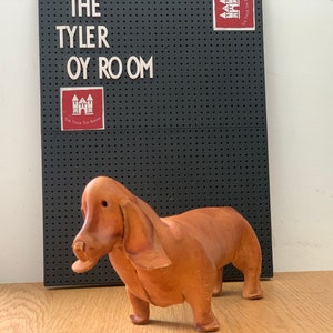 May include: A brown leather dachshund figurine. The figurine is standing on a wooden surface in front of a black pegboard with the text "The Tyler Toy Room" on it.