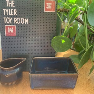 May include: Two blue ceramic dishes, one a small jug and the other a rectangular serving dish, on a wooden surface. The background is a black pegboard with the words "The Tyler Toy Room" written in white.