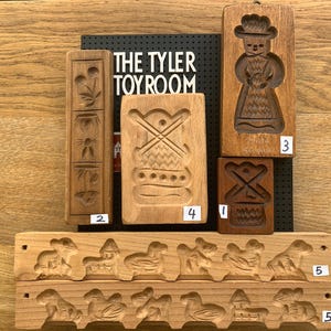 May include: A set of nine wooden cookie cutters, featuring various designs including a gingerbread person, a windmill, and a variety of animals. The cutters are arranged on a wooden surface with a black background.