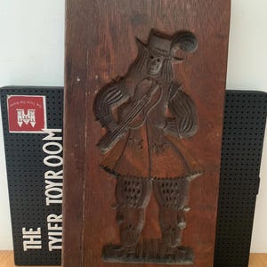 Pode incluir: Uma escultura em madeira vintage, castanho escuro, de uma figura em trajes de época, segurando um instrumento de cordas. A escultura é montada em uma placa de madeira retangular. A figura usa um chapéu com uma pena. O fundo inclui um suporte de livros preto com o texto "THE TYLER TOY ROOM".