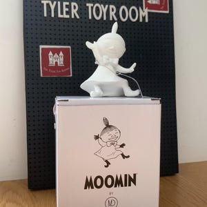May include: A white Moomin character figurine sits atop a box with the word "MOOMIN" printed on it. The box also features a cartoon illustration of the character. Behind the box is a black pegboard with the words "THE TYLER TOYROOM".