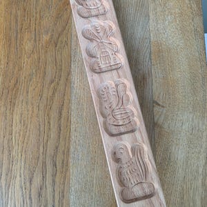 May include: A wooden springerle cookie mold with carved designs of a bird, windmill, swan, and other figures. The rectangular mold is made of light-colored wood and is angled on a wooden surface.