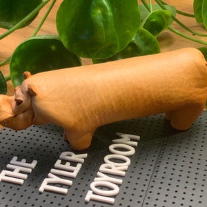 May include: A tan leather hippopotamus figurine. The figurine is standing on a black surface with white letters that read "The Tyler Toyroom".