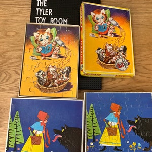 May include: Four vintage jigsaw puzzles featuring a cat, a girl in a red hood, and a wolf. The puzzles are on a wooden surface. The cat puzzle has a yellow background with a cat reading a book and kittens in a basket. The girl in a red hood puzzle has a blue background with a girl in a red hood and a wolf. The wolf puzzle has a blue background with a wolf and a girl in a red hood. The cat puzzle has the text "Jigsaw Puzzle" on it.