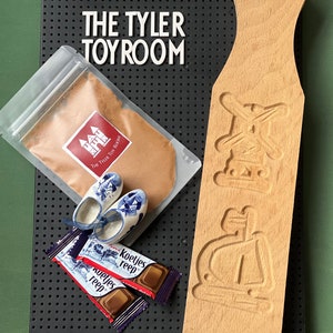 May include: A wooden cookie mold with two designs, a windmill and a ship, along with a bag of cookie dough mix, two small blue and white clogs, and two chocolate bars with the text "koekjes reep" on the wrappers. The text "The Tyler Toy Room" is visible on the bag of cookie dough mix.