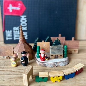 May include: Wooden Christmas village scene with a church, houses, Santa Claus, an angel, and a train. The scene is set on a wooden surface, with a flag and text in the background.