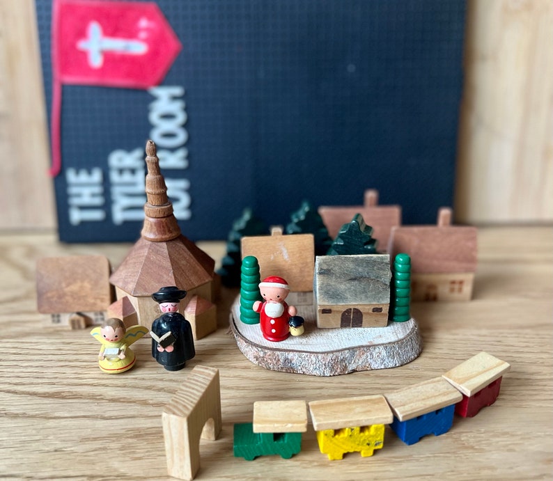 May include: Wooden Christmas village scene with a church, houses, Santa Claus, an angel, and a train. The scene is set on a wooden surface, with a flag and text in the background.