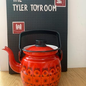 Puede incluir: Una tetera vintage de esmalte rojo con asa y tapa negras. La tetera presenta un estampado floral en tonos naranja y negro. El fondo incluye un letrero negro que dice "THE TYLER TOYROOM".