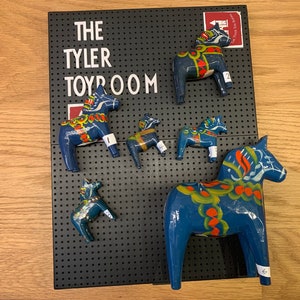 May include: A collection of six blue and white painted wooden horses, known as Dala horses, displayed on a black pegboard. The horses are of varying sizes and feature traditional Swedish folk art designs. The pegboard has the text "THE TYLER TOYROOM" printed on it.