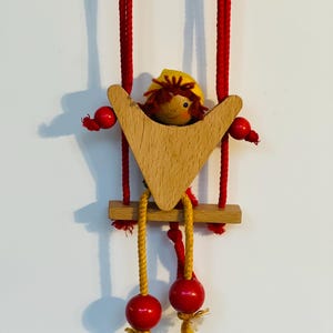 May include: A wooden toy with a red-haired figure sitting in a swing. The swing is made of wood and has red beads on the strings. The figure is wearing a yellow hat.