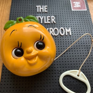 May include: A vintage orange-shaped wall hanging with a smiling face, black eyes, and green leaves. It has a string and a white plastic handle. The background includes the text "THE TYLER ROOM" on a black surface.