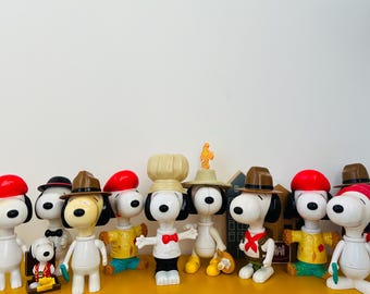 May include: A group of ten Snoopy figurines, each wearing a different hat and outfit. The figurines are all white with black noses and eyes. The hats include a chef's hat, a cowboy hat, a baseball cap, a beret, and a fedora. The outfits include a chef's uniform, a cowboy outfit, a baseball uniform, a beret, and a fedora.