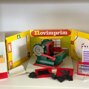 May include: A vintage Novimprim printing machine toy set. The set includes a green and red printing press, letter blocks, and a box with the text "Novimprim MACHINE A IMPRIMER". The box features a child using the machine.