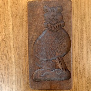 May include: A vintage, rectangular wooden mould featuring a carved design of a bear-like figure. The bear has a round head with small ears and a detailed body. The mould is a dark brown colour and appears well-worn.