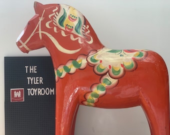 Puede incluir: Un caballo de balancín de madera rojo tradicional con detalles pintados en blanco, verde y amarillo. El caballo tiene una silla de montar y una brida.
