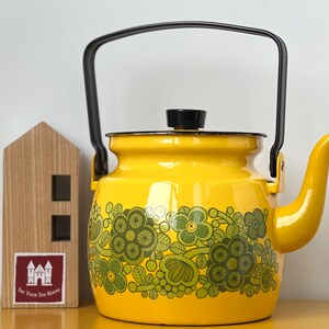 Puede incluir: Una tetera de esmalte amarillo brillante con un patrón floral verde oscuro alrededor de la base, un asa negra y un pomo de tapa negro. Junto a la tetera hay un pequeño bloque de madera con forma de casa y una etiqueta roja The Tiger Toy Room.
