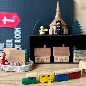 May include: Wooden toy village scene featuring a church, houses, and a train. The scene includes a church with a steeple, houses with brown roofs, and a colorful train. A banner with a cross and the text "THE TOY ROOM" is in the background.