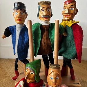 Könnte beinhalten: Eine Sammlung von fünf Handpuppen mit geschnitzten Holzköpfen und Filzkleidung. Drei Puppen stehen auf Holzstäben, während zwei kleinere Puppen davor platziert sind. Die Figuren haben bemalte Gesichtsdetails und farbenfrohe Stoffkleidung.