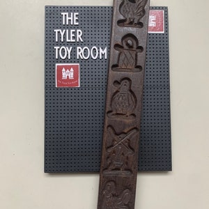 May include: A wooden cookie cutter with five different designs: a rooster, a hat, a bird, a windmill, and a bird. The cookie cutter is brown and has a worn look. The cookie cutter is on a black background with a white label that says "The Tyler Toy Room".