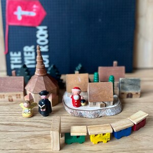 May include: A wooden toy village scene featuring a church, houses, and figures, including an angel and Santa Claus. The scene includes a train with colorful cars. A sign in the background reads "THE TYLER ROOM" with a cross.