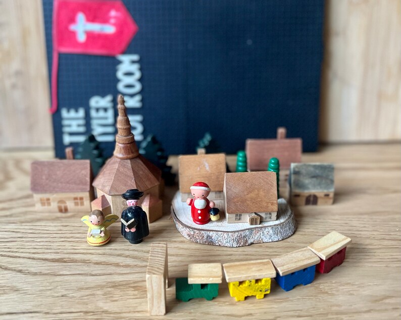 May include: A wooden toy village scene featuring a church, houses, and figures, including an angel and Santa Claus. The scene includes a train with colorful cars. A sign in the background reads "THE TYLER ROOM" with a cross.