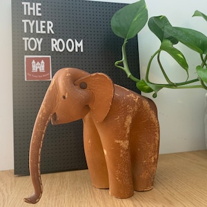 May include: A brown leather elephant figurine with a long trunk and a textured surface. The elephant is standing on a wooden surface.