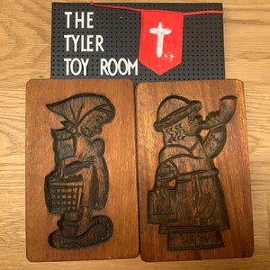 May include: Two wooden cookie cutters, one with a woman holding a basket and the other with a man playing a horn. The cookie cutters are both brown and have a rustic look.